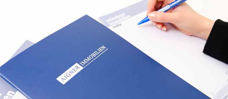 Blaue Mappe mit dem Aigner Immobilien Schriftzug und einer Hand, die mit einem Aigner-Kugelschreiber schreibt