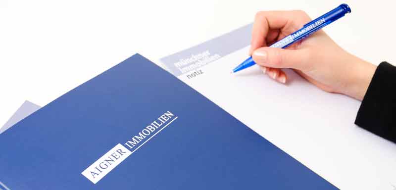 FAQ-Immobilien_800x384px Eine Frauenhand, die auf einem Notizblock schreibt, der neben einer Mappe von Aigner Immobilien liegt.