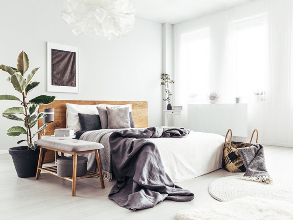 Gemütliches , helles Schlafzimmer mit einem Holzbett, mit weißen und grauen Kissen und Decken, sowie grauen Dekoelementen und Pflanzen