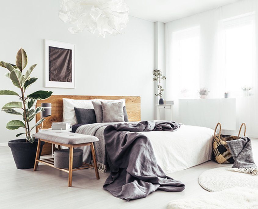 Gemütliches , helles Schlafzimmer mit einem Holzbett, mit weißen und grauen Kissen und Decken, sowie grauen Dekoelementen und Pflanzen