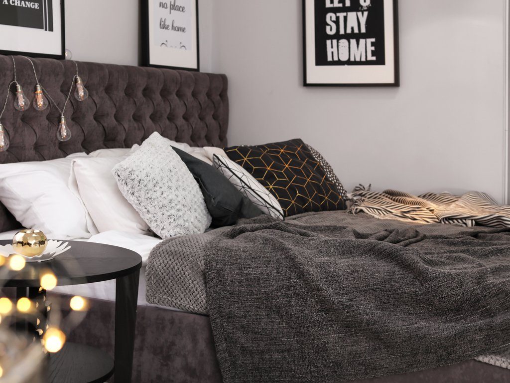 Dunkelgraues Bett mit hellen und dunklen Dekokissen. Eine Lichterkette über dem Kopfteil und schwarz, weiße Bilder an der Wand