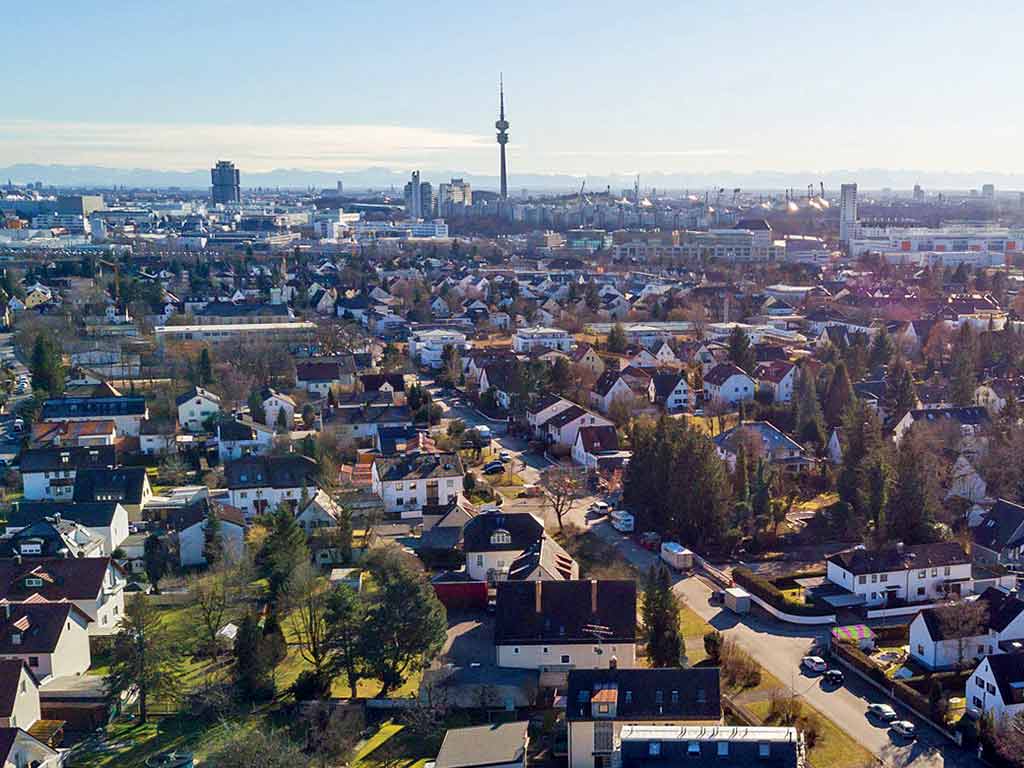 Luftbild eines Wohngebiets in München mit Einfamilienhäusern, viel Grün und dem Olympiaturm im Hintergrund, unter klarem blauem Himmel.