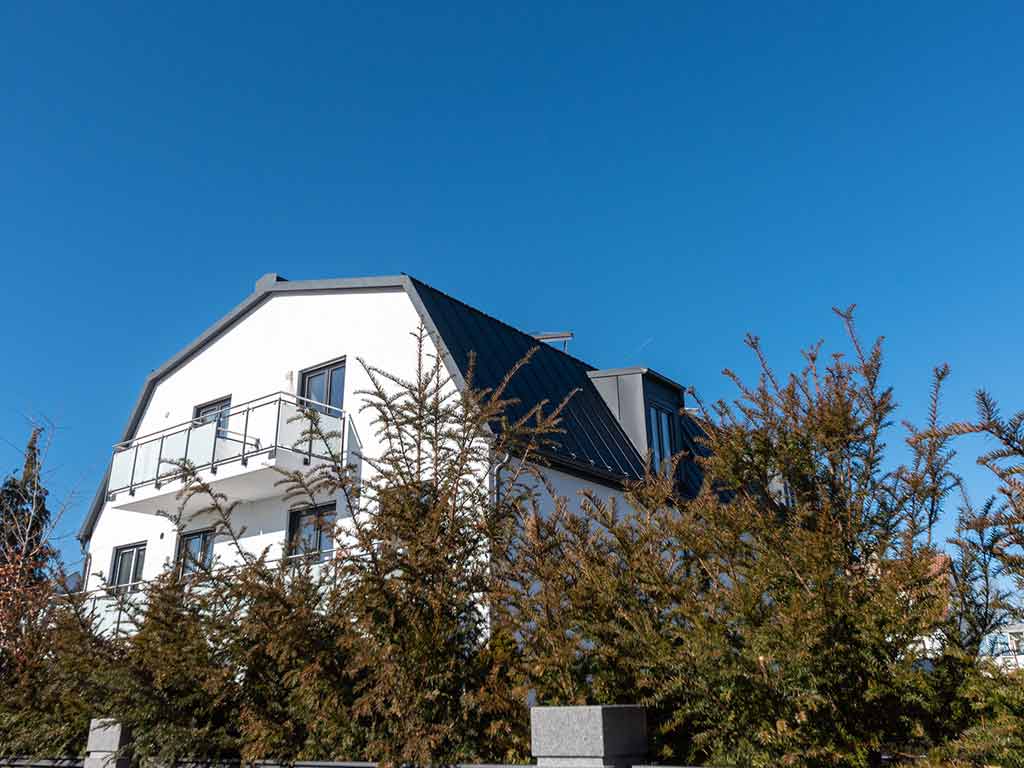 Außenansicht eines weißen Mehrfamilienhauses mit dunklem Dach, Balkonen und dichter Begrünung im Vordergrund unter strahlend blauem Himmel.
