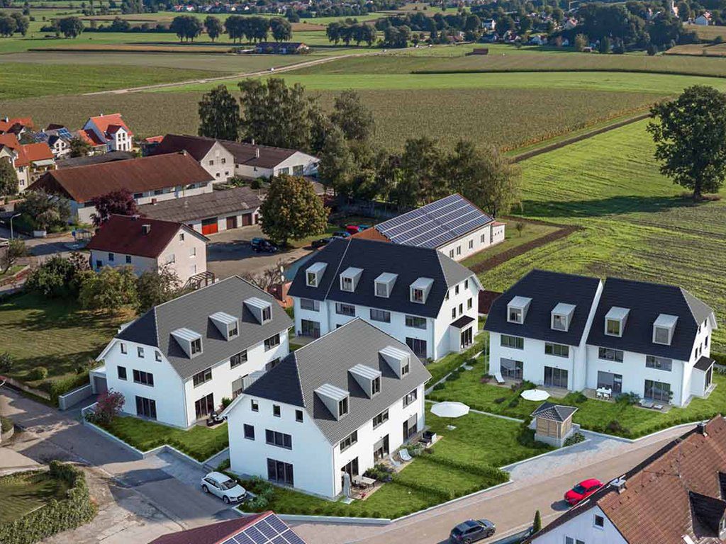 Luftaufnahme einer Neubau-Wohnsiedlung mit mehreren weißen Mehrfamilienhäusern in moderner Architektur. Grünflächen, Terrassen und Solarpaneele im Hintergrund. Ländliche Umgebung mit Feldern und Bauernhäusern.