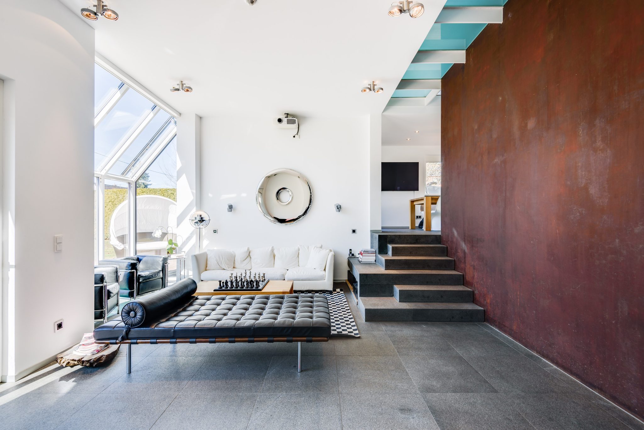 Stilvoll eingerichtetes Wohnzimmer mit hoher Decke, großen Glasfenstern und minimalistischen Möbeln. Eine schwarze Liege, ein weißes Sofa und ein Schachbrett-Tisch prägen den Raum. Eine markante rote Wand und moderne Architekturdetails verleihen dem Interieur einen Loft-Charakter.
