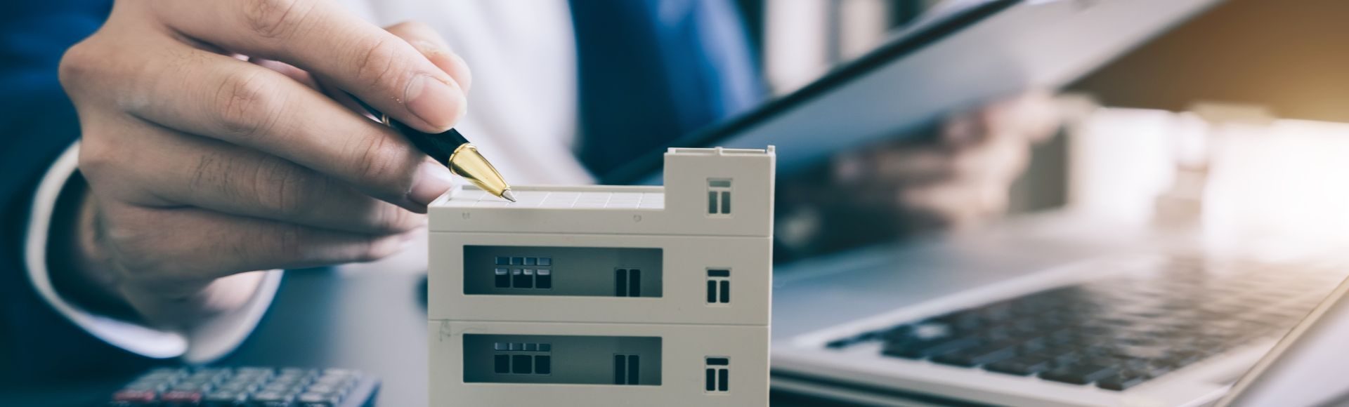 Notverkauf - Aigner Immobilien Hand schreibt mit einem Kugelschreiber auf einem Hausmodell, daneben liegt eine Tastatur