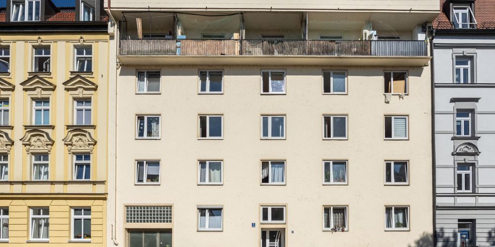 Fassade eines einfachen Mehrfamilienhauses mit großen Fenstern und einem durchgehenden Balkon im obersten Stockwerk