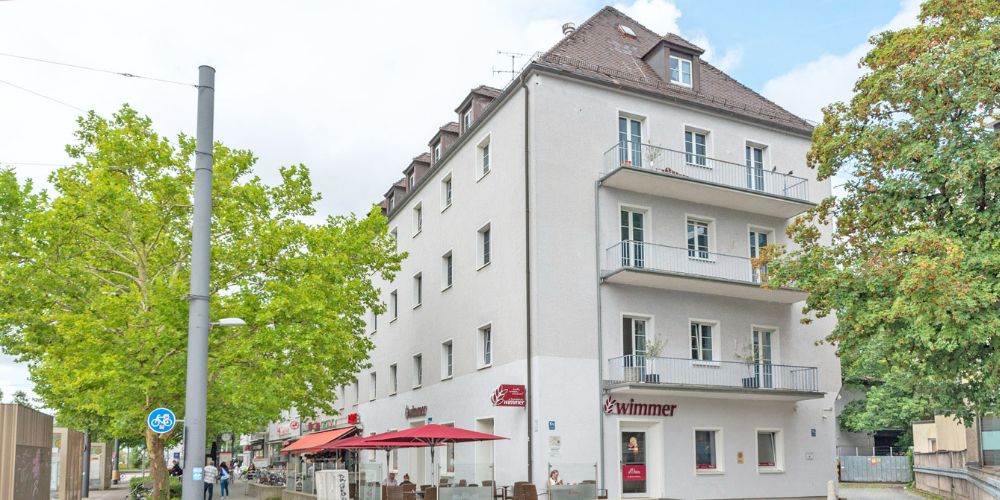 Eckgebäude eines Mehrfamilienhauses mit weißer Fassade, Balkonen und Geschäften, im Erdgeschoss eine Bäckerei