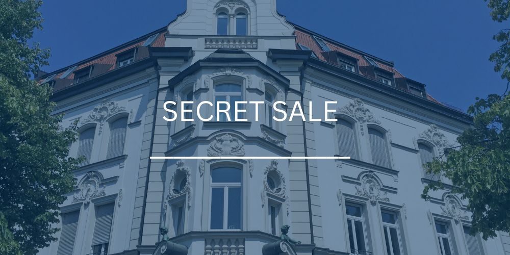 Detailreiche Fassade eines prachtvollen Jugendstil-Altbaugebäudes mit Dachgauben in München, eingefasst von grünen Bäumen, mit dem Schriftzug „Secret Sale“.