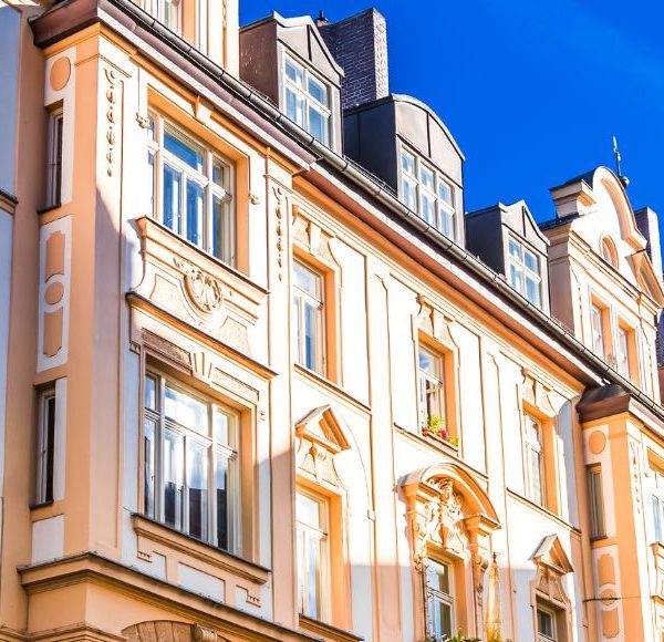 Historische Altbau-Fassade in München mit kunstvollen Verzierungen und großen Fenstern, beleuchtet von der Sonne vor einem strahlend blauen Himmel.