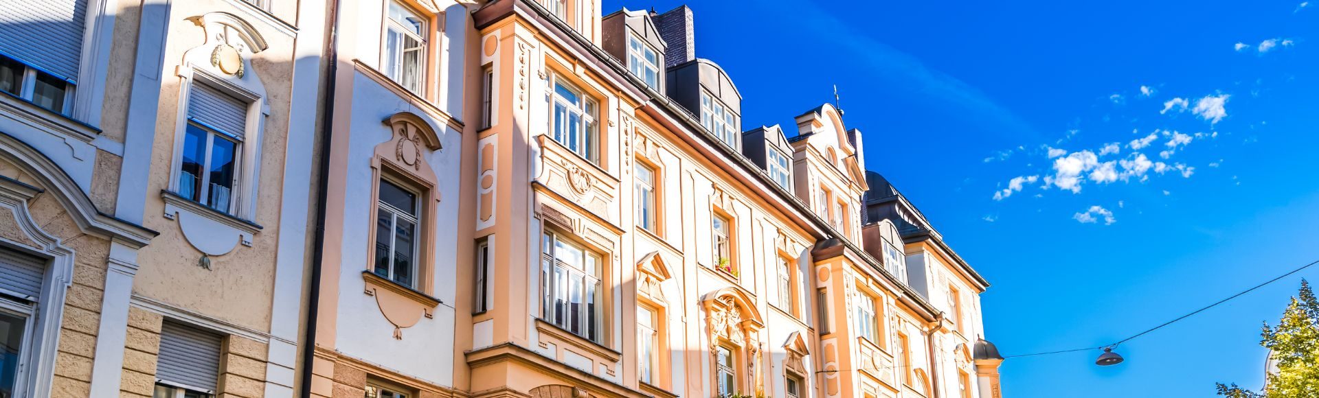 Historische Altbau-Fassade in München mit kunstvollen Verzierungen und großen Fenstern, beleuchtet von der Sonne vor einem strahlend blauen Himmel.