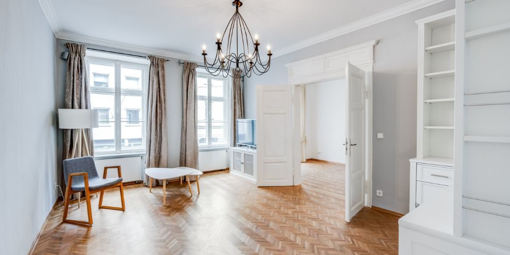 Wohnzimmer einer Altbauwohnung mit großen Fenstern, hellem Parkettboden, stilvollem Kronleuchter und klassischen Doppeltüren zur nächsten Wohnfläche.