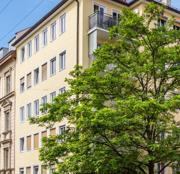 Altbau- und Neubau-Mehrfamilienhäuser in gepflegter Innenstadtlage mit Straßenbahnleitungen und grünem Straßenbaum unter blauem Himmel