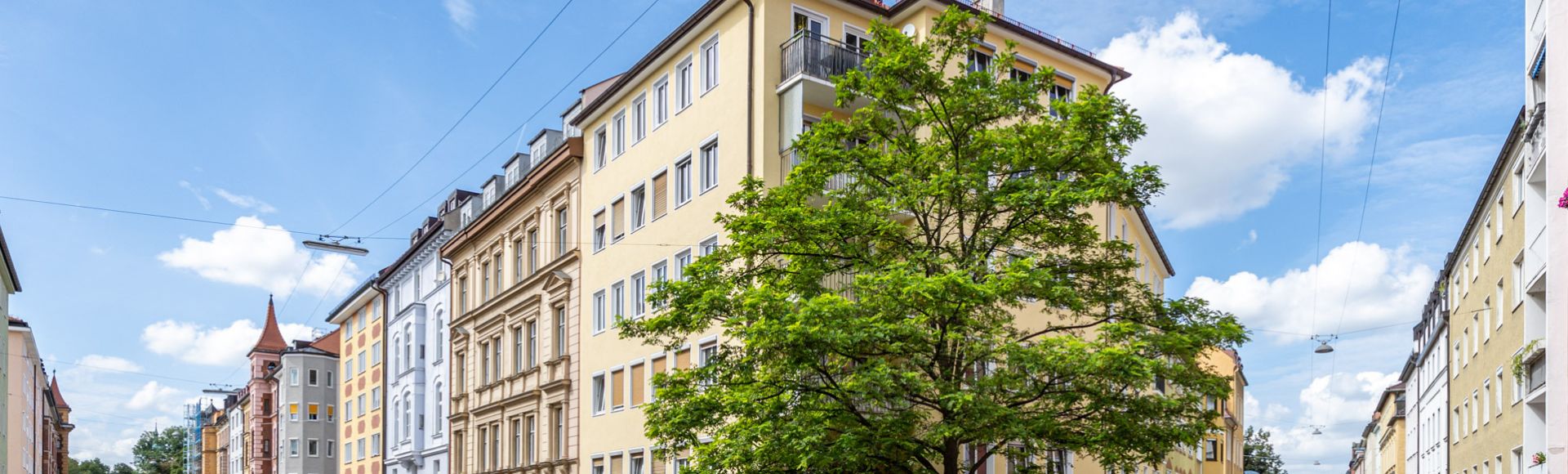 Altbau- und Neubau-Mehrfamilienhäuser in gepflegter Innenstadtlage mit Straßenbahnleitungen und grünem Straßenbaum unter blauem Himmel