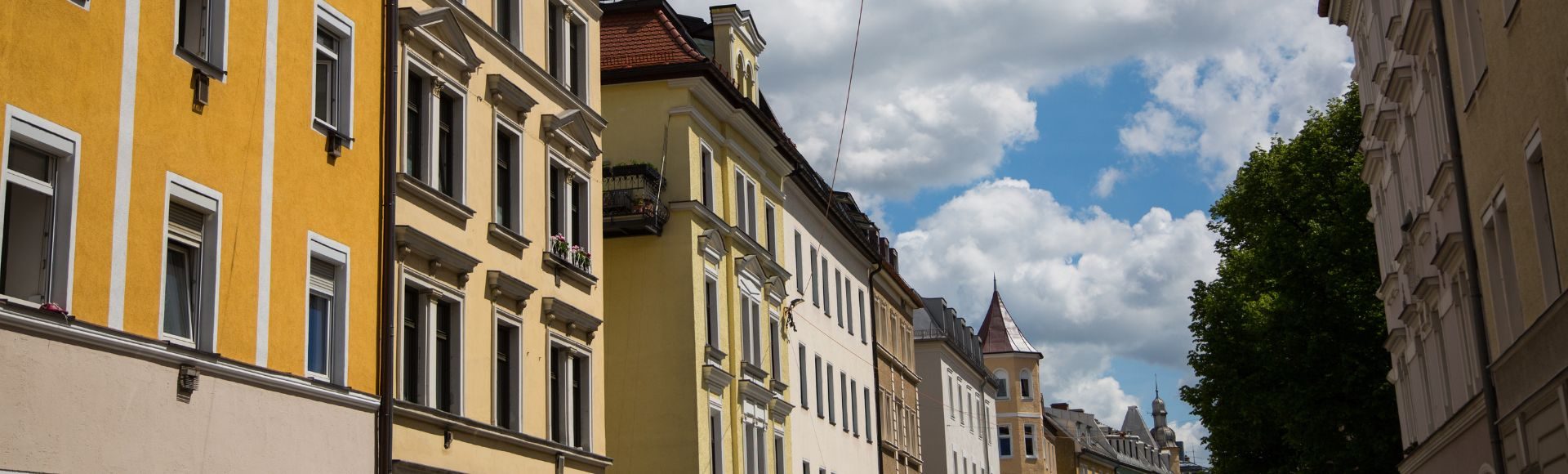 Historische Wohnhäuser mit bunten Fassaden in einer ruhigen Straße in München unter blauem Himmel mit Wolken