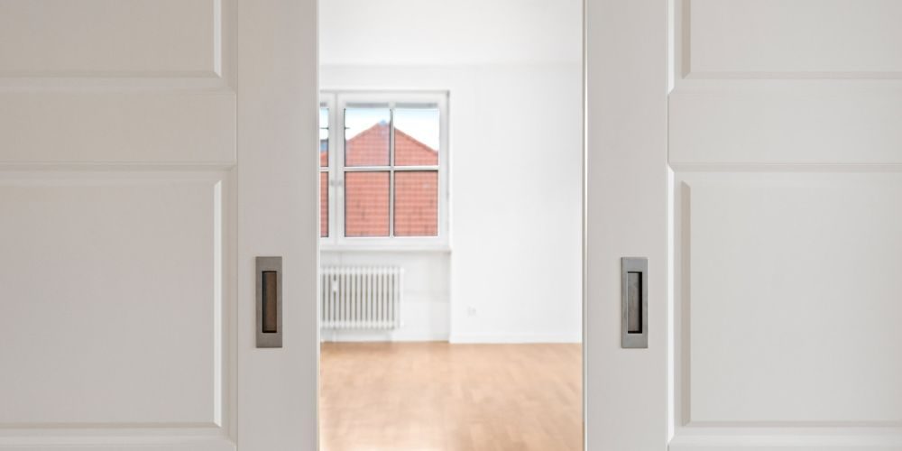 Offene weiße Schiebetüren mit Blick auf einen hellen Raum mit Parkettboden und großem Fenster in einer Münchner Altbauwohnung
