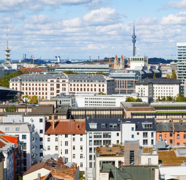Luftaufnahme von München mit Blick auf den Olympiaturm, historische Dächer und moderne Bürogebäude bei blauem Himmel