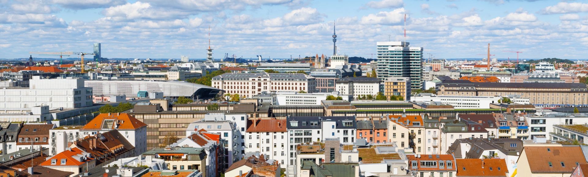 Luftaufnahme von München mit Blick auf den Olympiaturm, historische Dächer und moderne Bürogebäude bei blauem Himmel