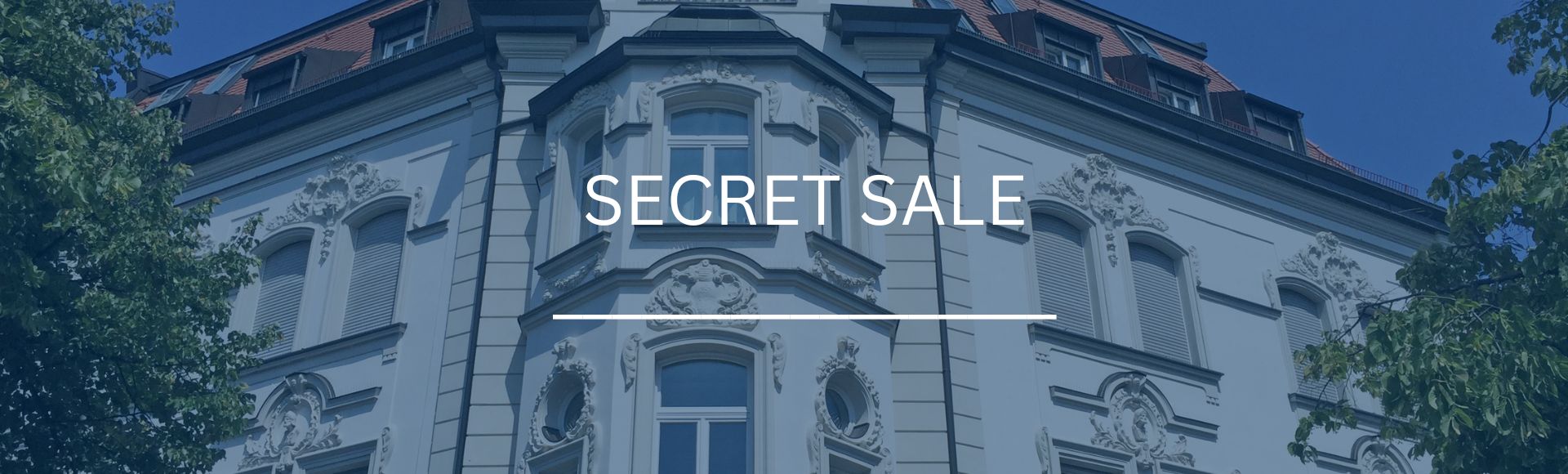 Detailreiche Fassade eines prachtvollen Jugendstil-Altbaugebäudes mit Dachgauben in München, eingefasst von grünen Bäumen, mit dem Schriftzug „Secret Sale“.
