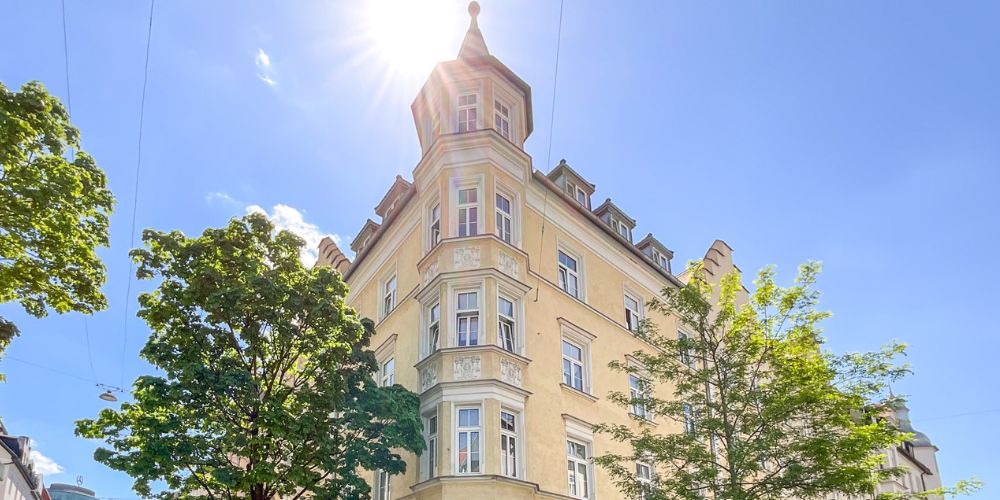 Historischer Altbau mit markantem Eckturm in München bei strahlendem Sonnenschein, umgeben von Bäumen und blauem Himmel