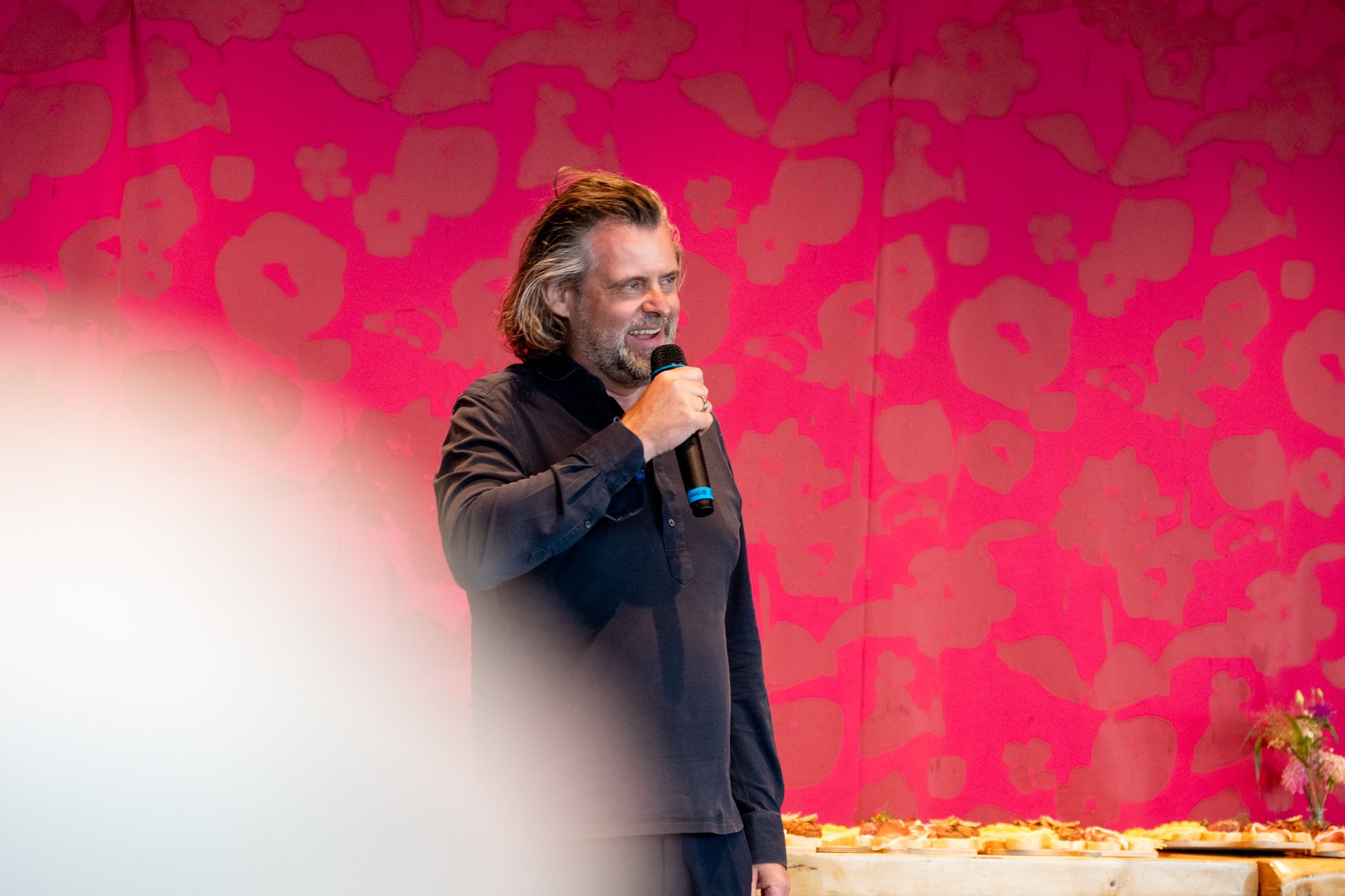 Thomas Aigner spricht mit Mikrofon vor pinker Wand beim Aigner Immobilien Sommerfest 2025