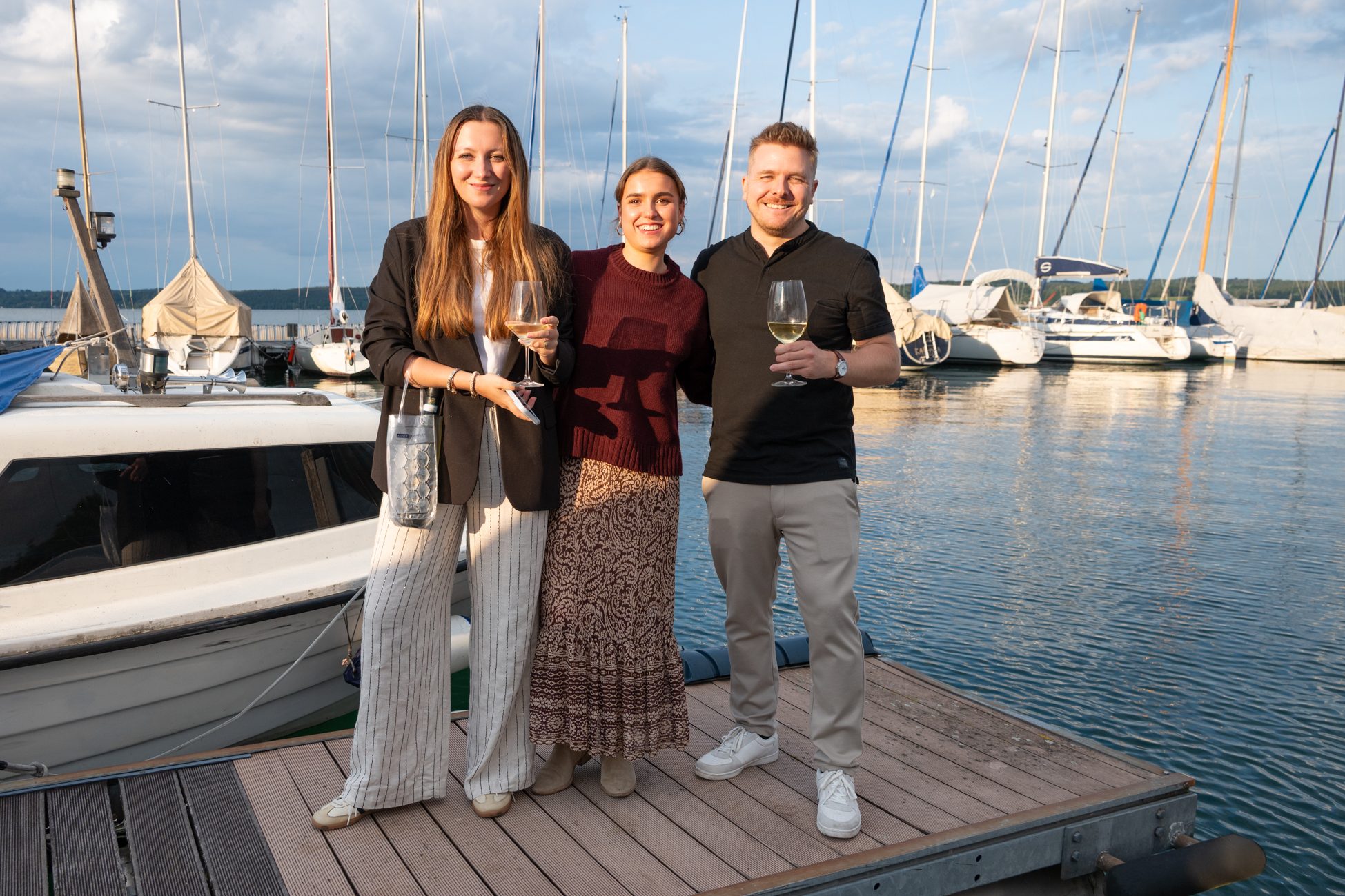 Drei Gäste mit Weingläsern am Bootssteg vor Segelbooten