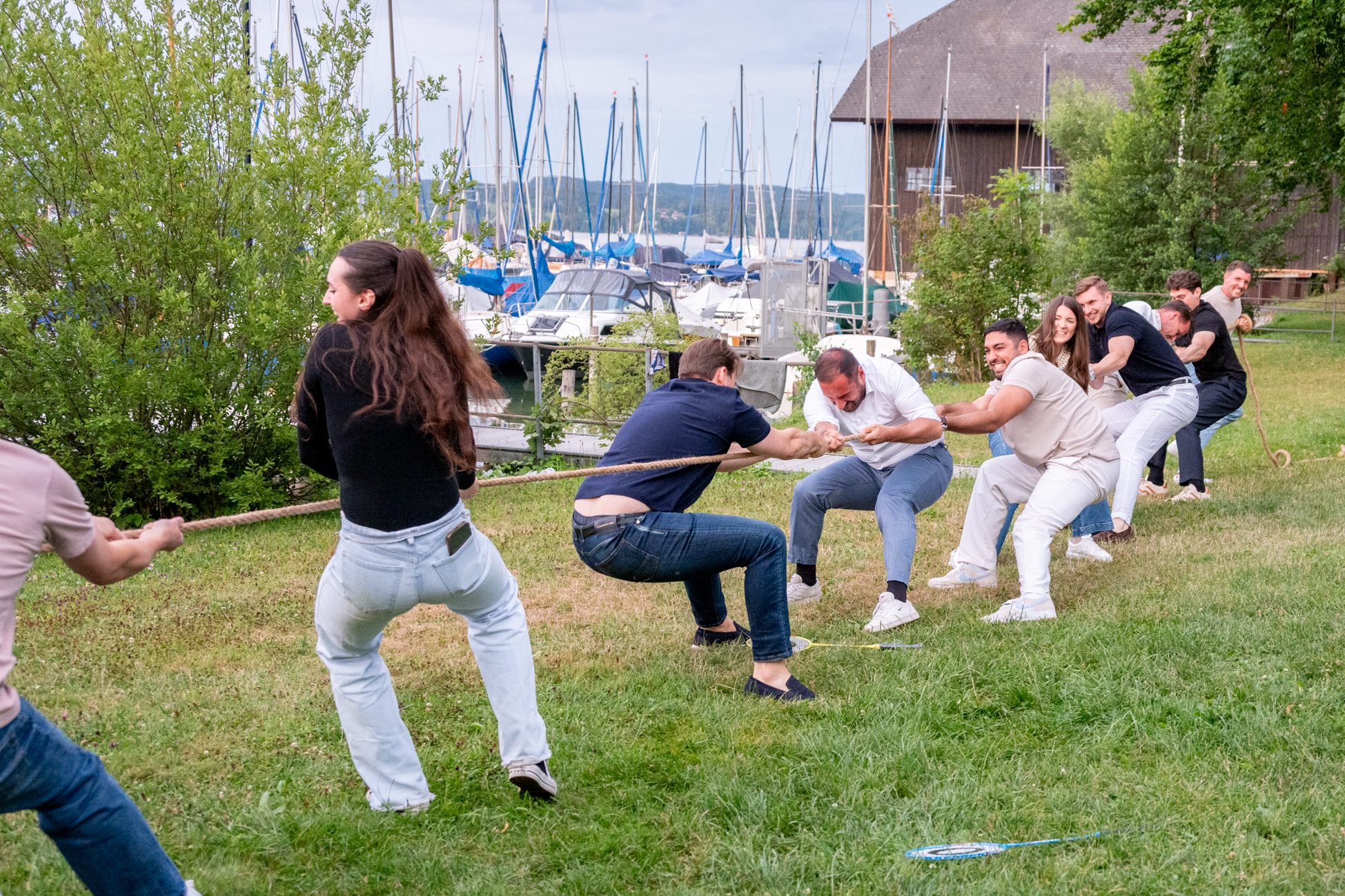 Gruppe junger Menschen beim Tauziehen auf einer Wiese mit Blick auf Segelboote