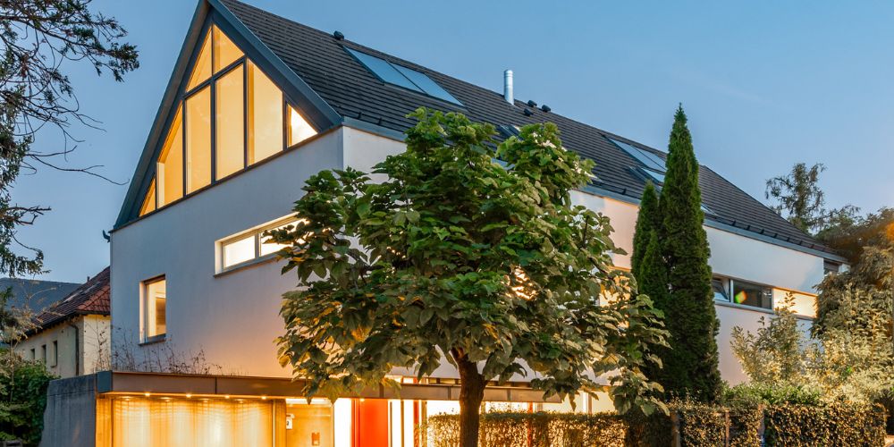 Modernes Einfamilienhaus in München (39379 ) Modernes Einfamilienhaus in München mit großer Glasfassade, Satteldach und beleuchtetem Eingangsbereich am Abend, umgeben von Bäumen und gepflegtem Garten
