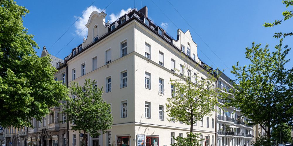 Helles, saniertes Altbau-Mehrfamilienhaus mit dekorativem Giebel und Ladengeschäften im Erdgeschoss in München, umgeben von grünen Bäumen und blauem Himmel