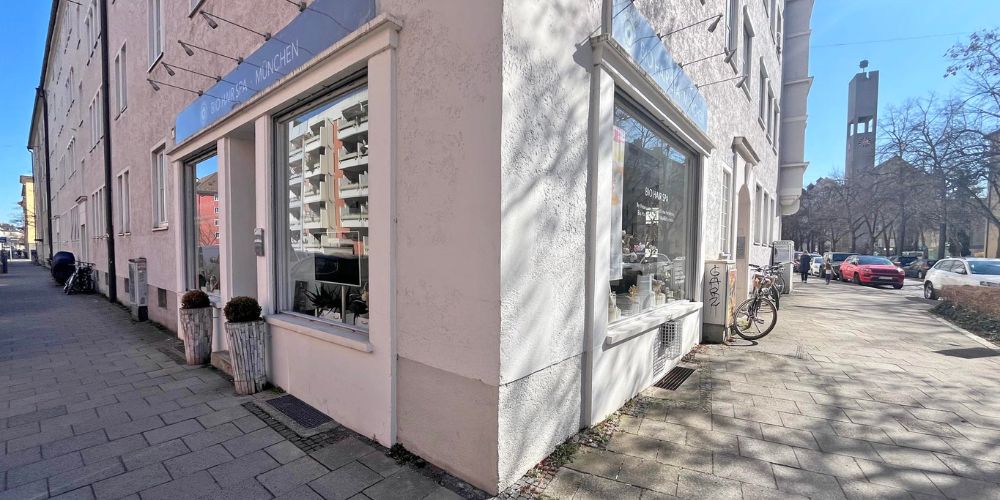 Eckhaus mit moderner Ladenfläche und großer Fensterfront in München, sonnige Straßenszene mit Wohngebäuden und Kirchturm im Hintergrund