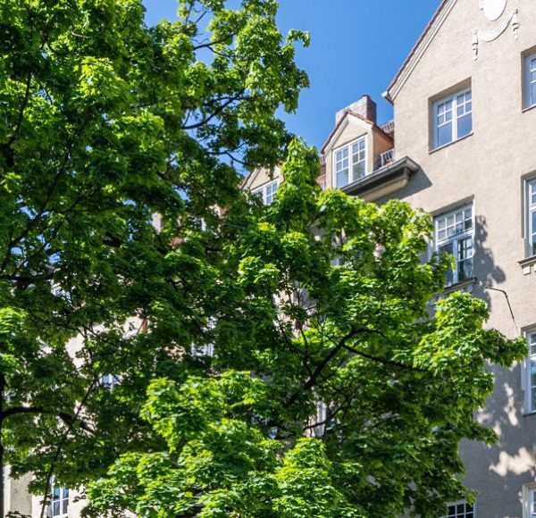 Helles Mehrfamilienhaus im Altbaustil in München, umgeben von grünen Bäumen und blauem Himmel an einem sonnigen Tag