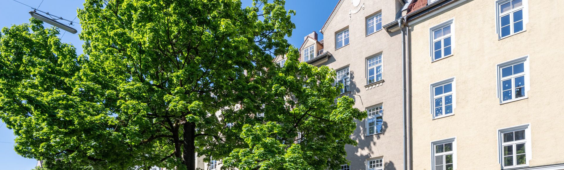 Helles Mehrfamilienhaus im Altbaustil in München, umgeben von grünen Bäumen und blauem Himmel an einem sonnigen Tag