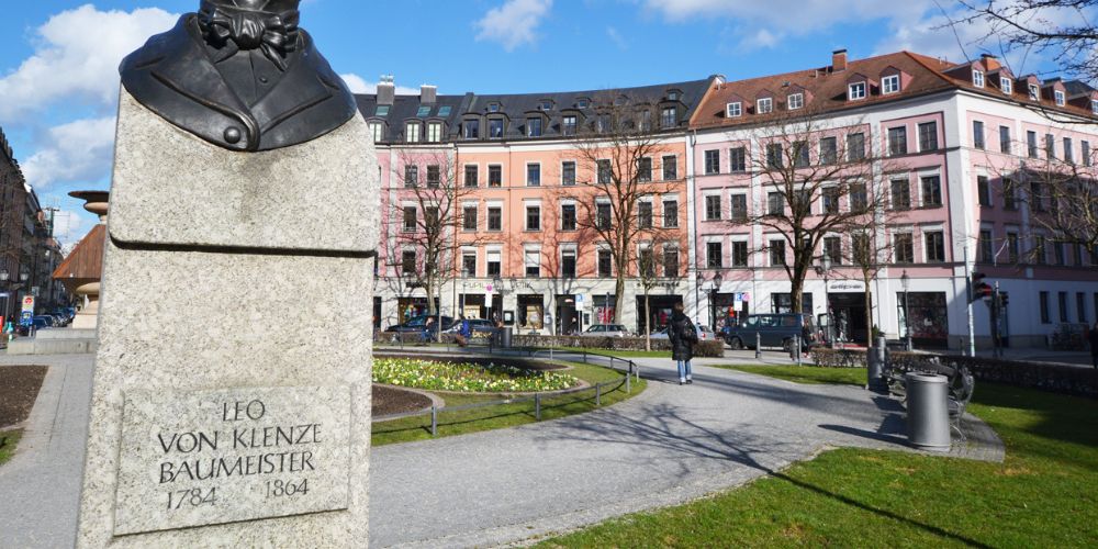 Gärtnerplatz in München mit Denkmal von Leo von Klenze im Vordergrund und restaurierten Altbaufassaden im Hintergrund bei sonnigem Wetter