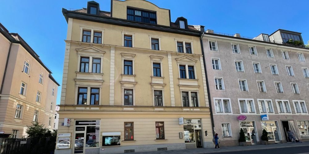 Straßenansicht eines gepflegten Altbauhauses in München mit Stuckfassade, hohen Fenstern und Ladengeschäften im Erdgeschoss
