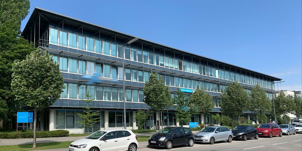 Außenansicht eines modernen Bürogebäudes in München mit großer Glasfassade, Bäumen und parkenden Autos entlang der Straße