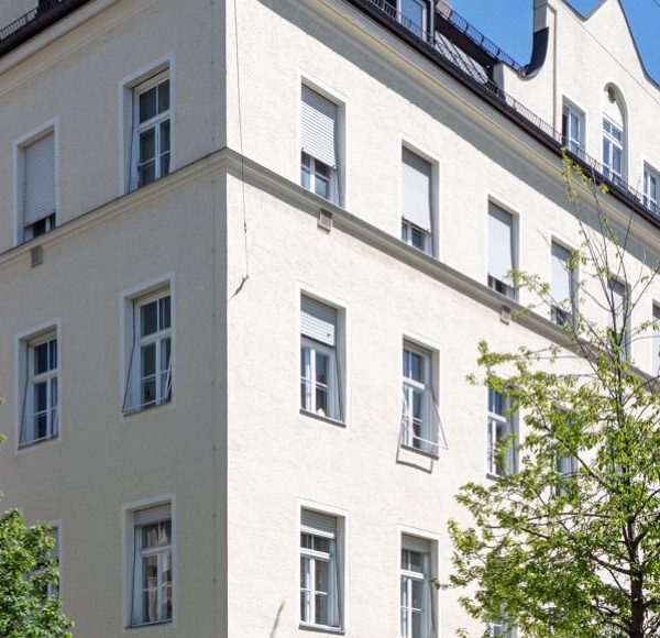 Helles Altbau-Mehrfamilienhaus in München mit weißen Fassaden und grünen Bäumen bei blauem Himmel