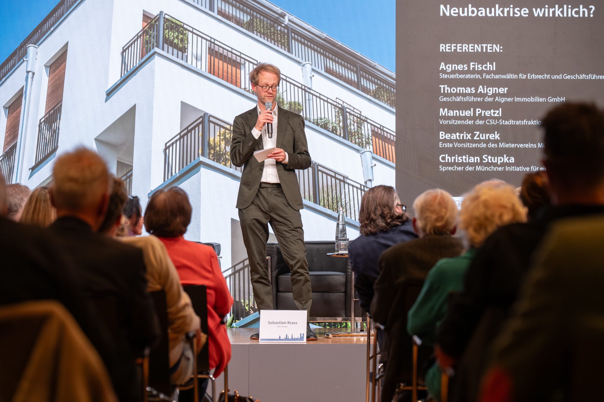 Moderator eröffnet Diskussionsrunde beim Immobilien Fokus Moderator auf Bühne beim Münchner Immobilien Fokus 2025 mit Publikum und Expertenpanel im Hintergrund