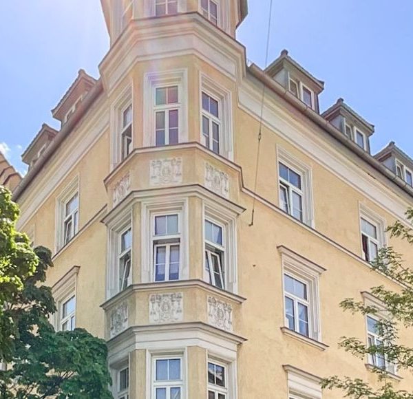 Historischer Altbau mit markantem Eckturm in München bei strahlendem Sonnenschein, umgeben von Bäumen und blauem Himmel