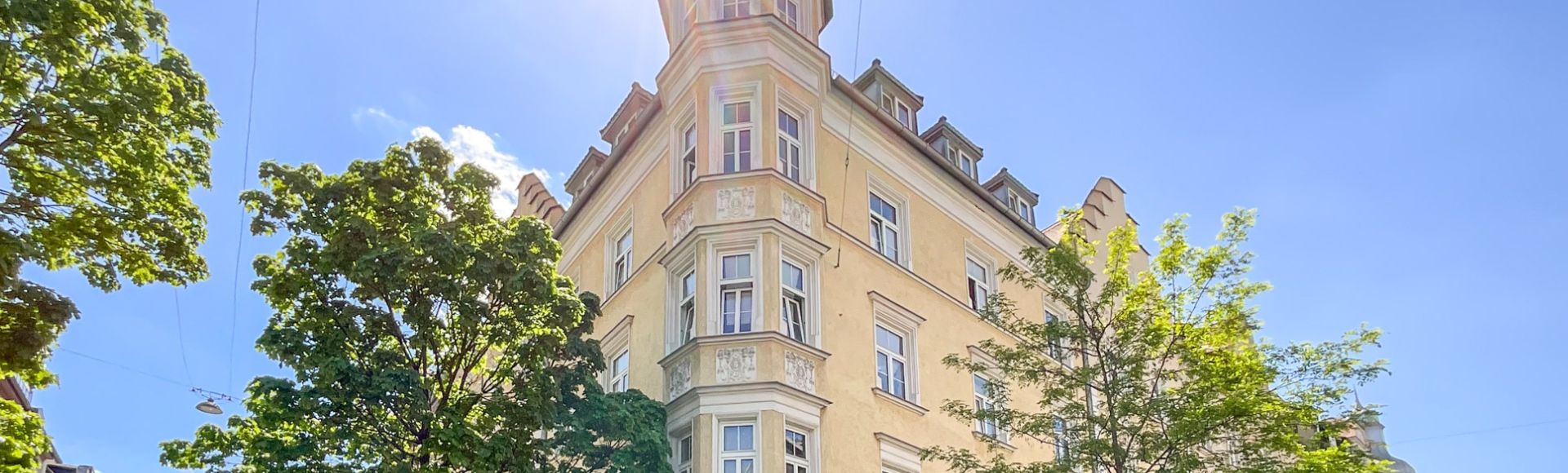 Historischer Altbau mit markantem Eckturm in München bei strahlendem Sonnenschein, umgeben von Bäumen und blauem Himmel