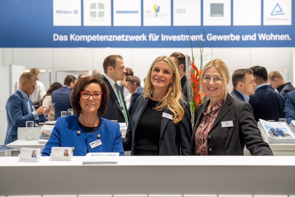 Expo Real 2025: 3 Mitarbeiterinnen von Aigner Immobilien fröhlich lachend am Messestand