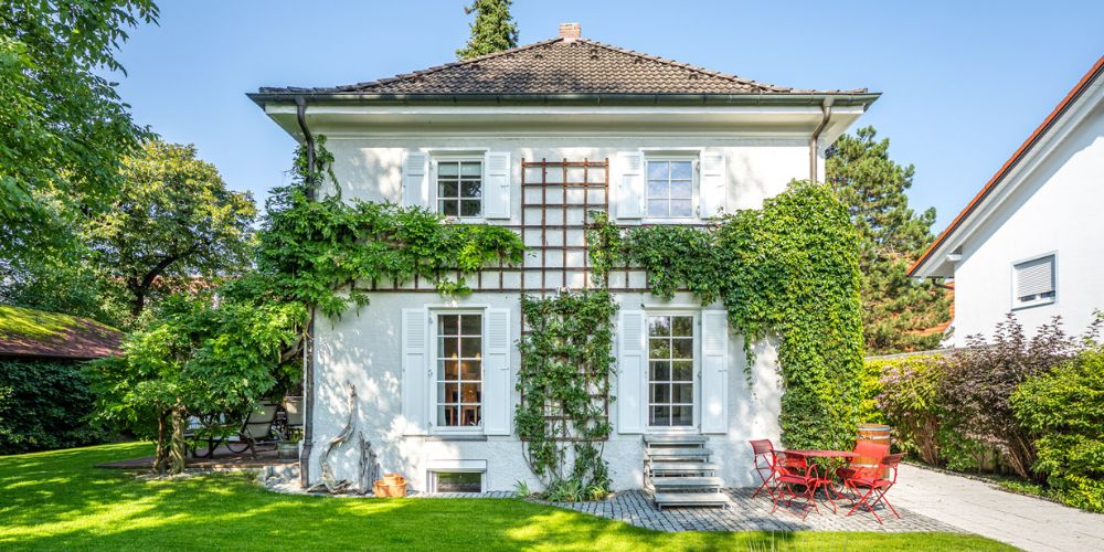 Exklusive Immobilie: Einfamilienhaus mit Garten (42331) Frontalansicht eines freistehenden Einfamilienhauses mit weißer Putzfassade, Fensterläden, Efeu und Rankgitter, kleiner Terrasse mit roter Sitzgruppe und gepflegtem Garten