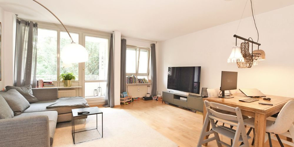Helles Wohnzimmer in München mit grauem Sofa, großem Fernseher, Holzesstisch mit Arbeitsplatz, bodentiefen Fenstern und hellem Parkettboden