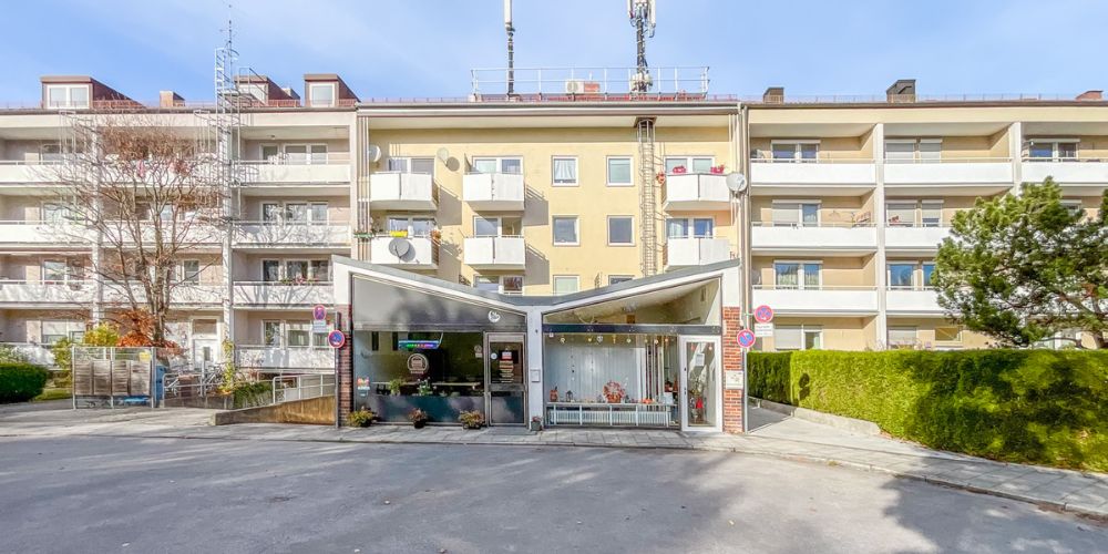 Wohn- und Geschäftshaus in München mit vorgelagertem Gewerbepavillon, Balkonen, Satellitenschüsseln und mehrstöckiger Wohnanlage im Hintergrund