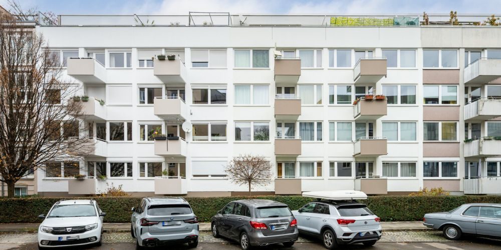 Mehrfamilienhaus in München mit weißer Fassade, mehreren Balkonen, Hecke im Vordergrund und parkenden Autos