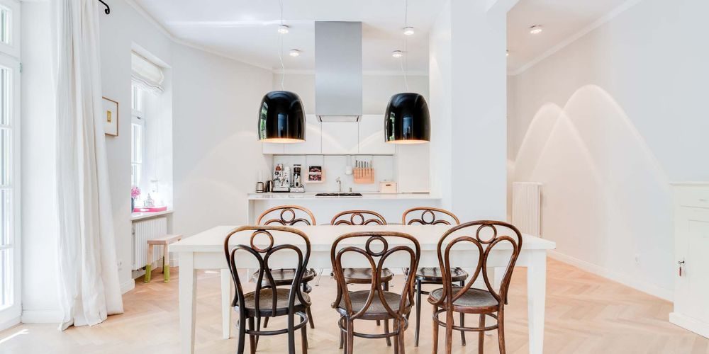 Eleganter Essbereich mit offener Küche in moderner Münchner Wohnung (29791 Stilvoller Essbereich einer Wohnung in München mit großem Esstisch, klassischen Holzstühlen, offener weißer Küche und modernen Pendelleuchten in hellem Ambiente