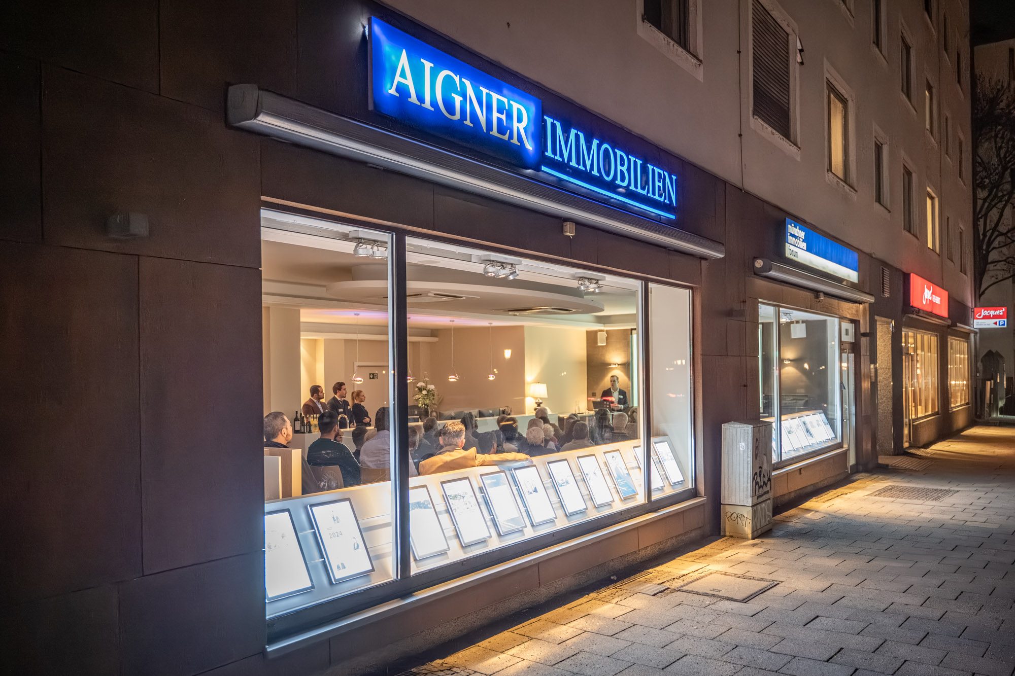 Außenansicht von Aigner Immobilien in München bei einer Abendveranstaltung mit Gästen im Schaufenster
