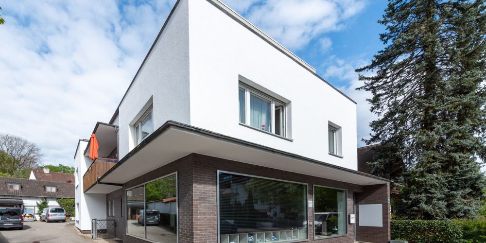 Moderne Wohn- und Gewerbeimmobilie mit weißer Fassade, großflächigen Schaufenstern und Balkon in urbaner Lage