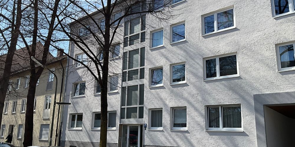 Helle Mehrfamilienhaus-Fassade in München mit modernen Fenstern, Eingangsbereich und ruhiger Wohnstraße mit Bäumen