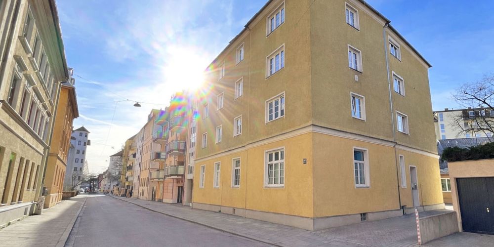 Vierstöckiges Wohnhaus an einer ruhigen Straße in München mit gelber Fassade, weißen Fensterrahmen, angrenzenden Gebäuden und Sonne, die von hinten auf die Fassade scheint.