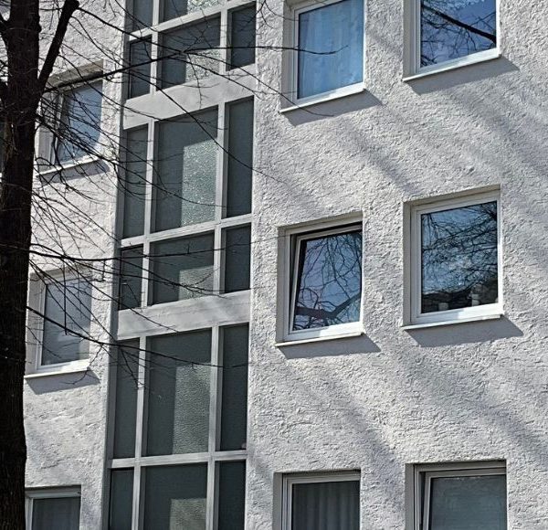 Außenansicht moderner Mehrfamilienhäuser in München bei Sonnenschein, weiße Fassaden, zahlreiche Fenster und Bäume im Vordergrund